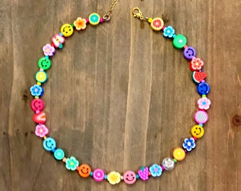 Op de afbeelding: Een kleurrijke kralenketting met een gouden ketting. De kralen zijn gevormd als smiley's, bloemen, fruit en andere leuke ontwerpen.