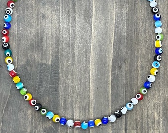 May include: A colourful necklace made of small glass beads with an evil eye design. The beads are in various colours, including blue, green, red, yellow, and black. The necklace is strung on a silver chain.