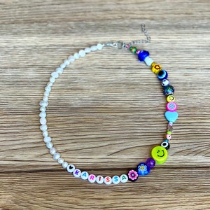 May include: A colorful beaded necklace with a mix of white pearls and vibrant beads. The necklace features the name "KARISSA" spelled out in letter beads, along with smiley faces, hearts, and flower designs. The necklace is on a wooden surface.