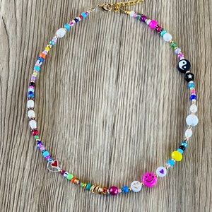 Op de afbeelding: Een kleurrijke kralenketting met verschillende bedels, waaronder een yin yang symbool, een smiley en een hart. De ketting is gemaakt van kleine, kleurrijke kralen en heeft een gouden sluiting.