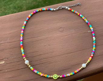 Op de afbeelding: Een kleurrijke kralenketting met een smiley-bedel en twee witte parelkraaltjes. De ketting is gemaakt met kleine, ronde kralen in verschillende kleuren, waaronder rood, oranje, geel, groen, blauw en paars.