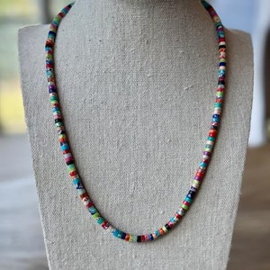 Op de afbeelding: Een kleurrijke kralenketting met een verscheidenheid aan kleine, gepatroneerde kralen in tinten rood, blauw, groen, geel en wit. De ketting wordt getoond op een witte, met stof beklede mannequin.