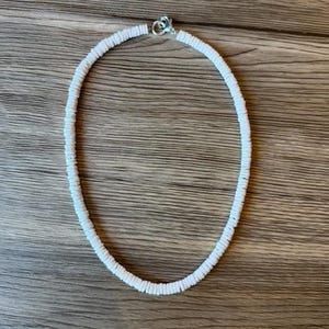 May include: White beaded necklace with a silver clasp. The necklace is made of small, round, white beads strung together. The necklace is displayed on a wooden surface.