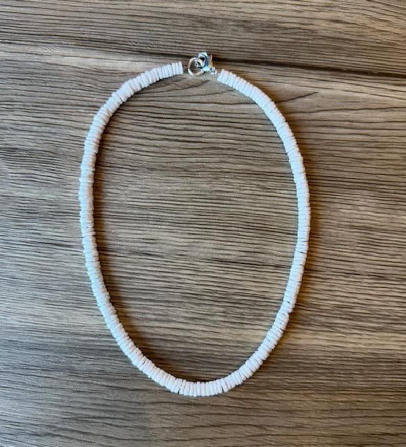 Op de afbeelding: Witte kralenketting met een zilveren sluiting. De ketting is gemaakt van kleine, ronde, witte kralen die aan elkaar zijn geregen. De ketting wordt getoond op een houten oppervlak.
