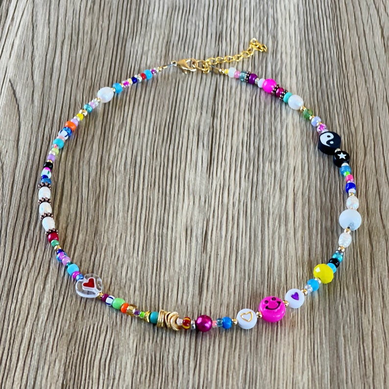 Op de afbeelding: Een kleurrijke kralenketting met verschillende bedels, waaronder harten, smiley-gezichten en een yin-yang symbool. De ketting is gemaakt met witte, roze, blauwe, gele, groene en rode kralen.