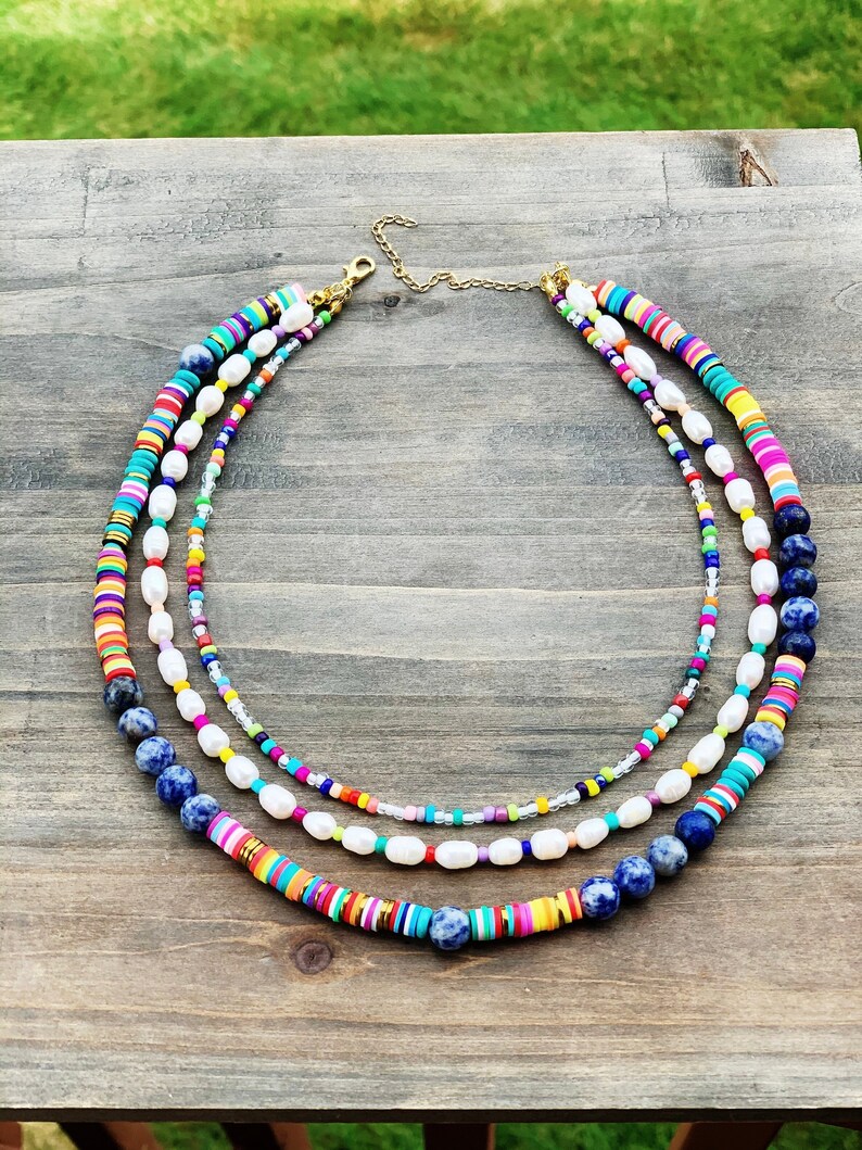 May include: A multi-layered necklace with colorful beads, white pearls, and blue stones. The necklace is made of different materials and has a gold clasp.