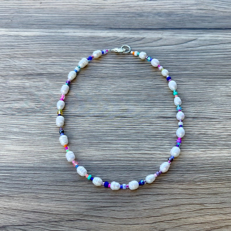May include: A white pearl necklace with colourful beads. The necklace is on a wooden surface.