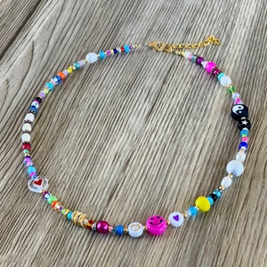 Op de afbeelding: Een kleurrijke kralenketting met een gouden kettingsluiting. De kralen zijn in verschillende vormen en maten, waaronder harten, smiley-gezichten en een yin-yang-symbool. De ketting ligt op een houten oppervlak.