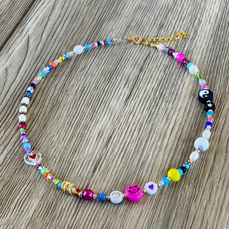 Op de afbeelding: Een kleurrijke kralenketting met een gouden kettingsluiting. De kralen zijn in verschillende vormen en maten, waaronder harten, smiley-gezichten en een yin-yang-symbool. De ketting ligt op een houten oppervlak.
