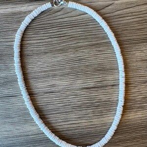 Op de afbeelding: Een witte kralenketting met een zilveren sluiting. De ketting is gemaakt van kleine, platte, ronde kralen die aan elkaar zijn geregen om een cirkelvorm te vormen. De ketting wordt gepresenteerd op een houten oppervlak.