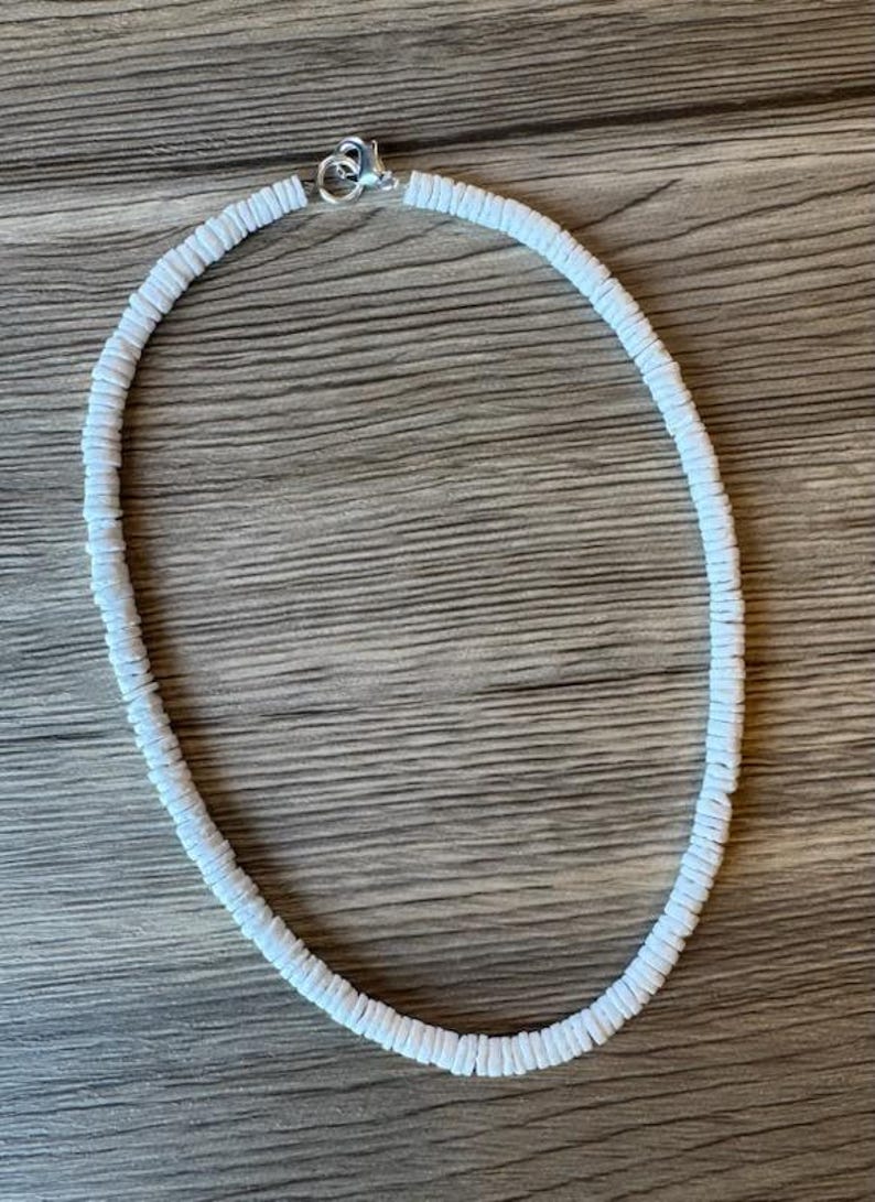 Op de afbeelding: Een witte kralenketting met een zilveren sluiting. De ketting is gemaakt van kleine, platte, ronde kralen die aan elkaar zijn geregen om een cirkelvorm te vormen. De ketting wordt gepresenteerd op een houten oppervlak.