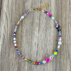 Op de afbeelding: Een kleurrijke kralenketting met verschillende bedels, waaronder een yin yang symbool, een smiley, een hartje en een ster. De ketting is gemaakt van witte, roze, blauwe, gele, groene en rode kralen.