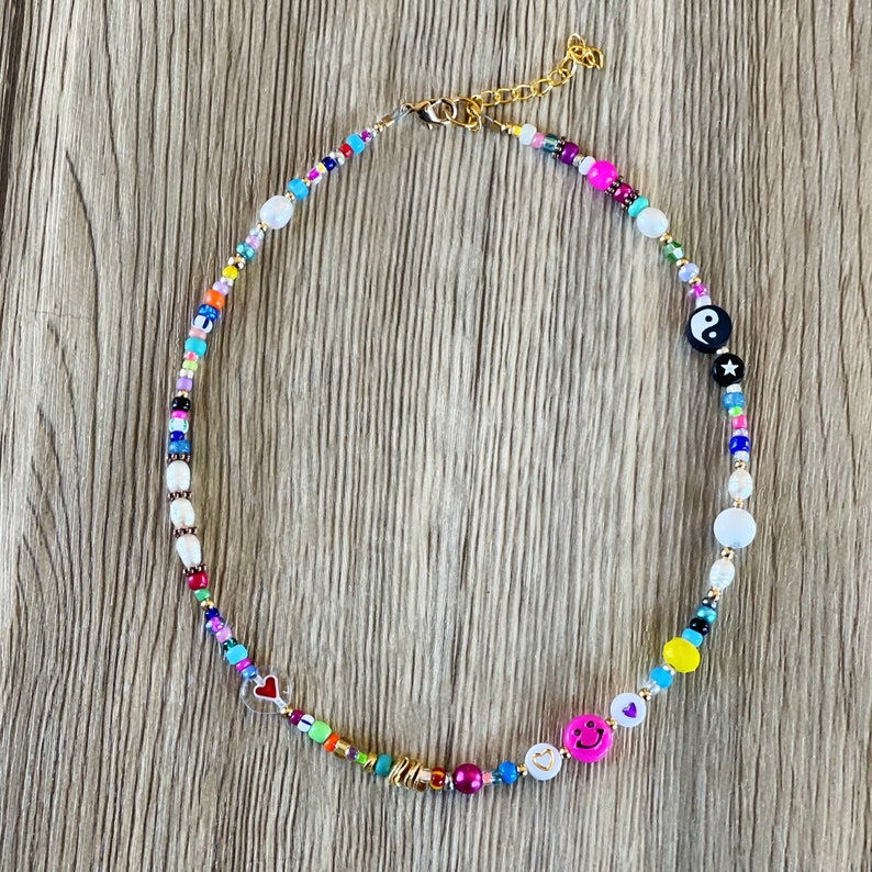Op de afbeelding: Een kleurrijke kralenketting met verschillende bedels, waaronder een yin yang symbool, een smiley, een hartje en een ster. De ketting is gemaakt van witte, roze, blauwe, gele, groene en rode kralen.