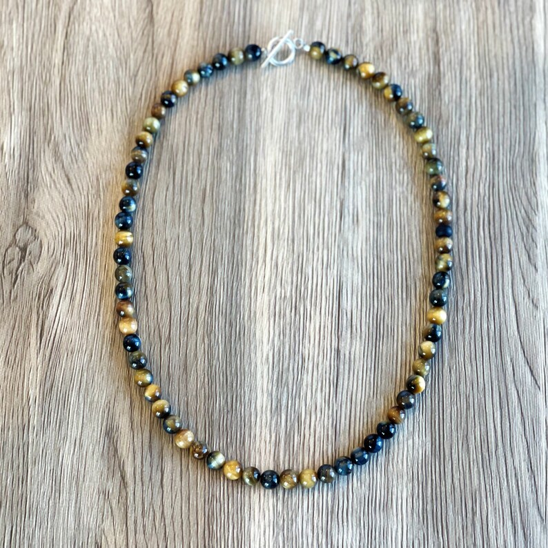 May include: A necklace made of tiger's eye beads in shades of gold, brown, and blue. The beads are round and have a smooth, polished finish. The necklace is secured with a silver clasp.