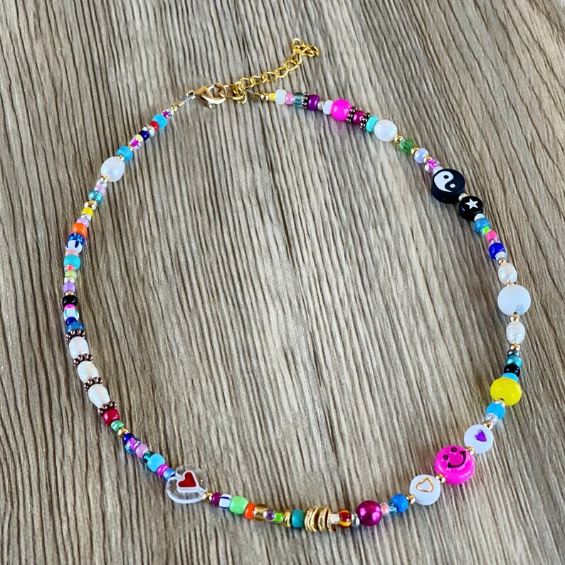 Op de afbeelding: Een kleurrijke kralenketting met verschillende bedels, waaronder een yin yang symbool, een hartje en een smiley. De ketting is gemaakt van witte, roze, blauwe, groene, gele en zwarte kralen.