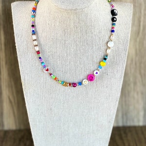 Op de afbeelding: Een kleurrijke kralenketting met verschillende bedels, waaronder harten, smiley-gezichten en een yin-yang symbool. De ketting is gemaakt van witte parels en gouden kralen.