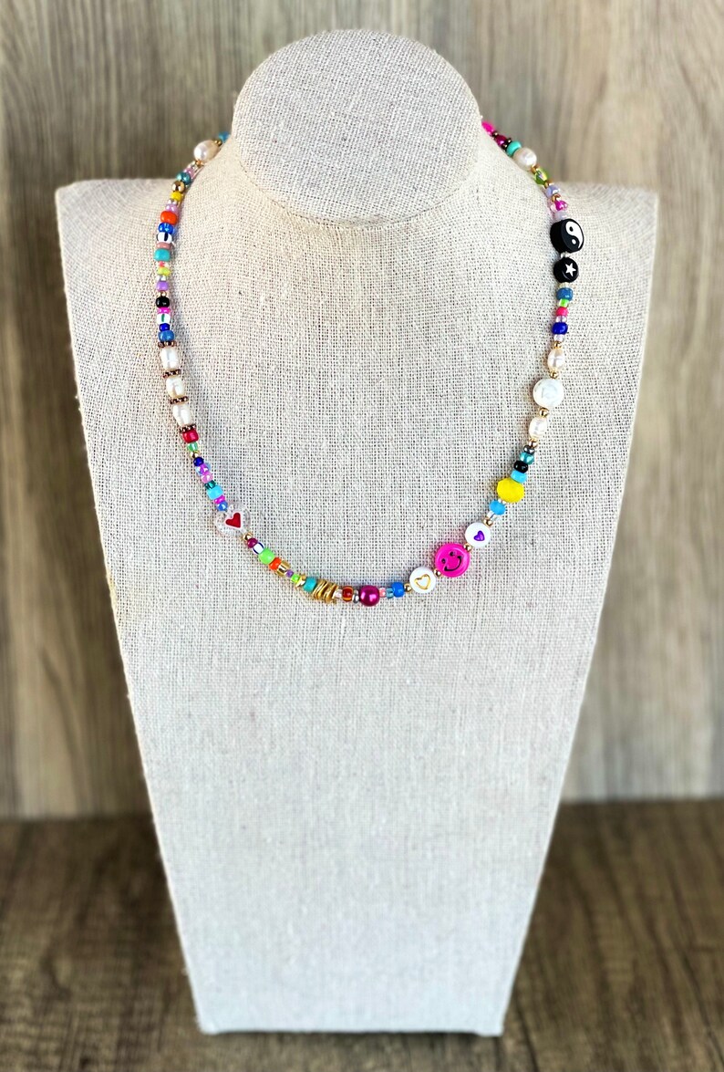 Op de afbeelding: Een kleurrijke kralenketting met verschillende bedels, waaronder harten, smiley-gezichten en een yin-yang symbool. De ketting is gemaakt van witte parels en gouden kralen.