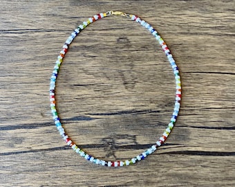May include: A colorful beaded necklace with a gold clasp. The beads are arranged in a rainbow pattern with white beads interspersed throughout.