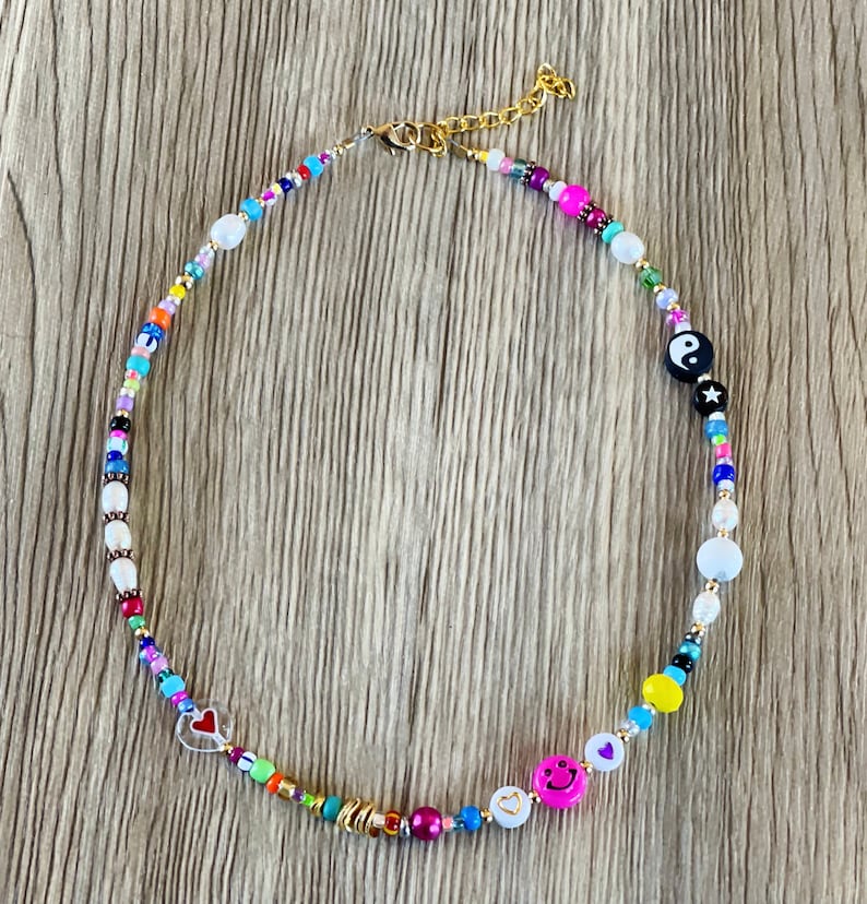 Op de afbeelding: Een kleurrijke kralenketting met verschillende bedels, waaronder een yin yang symbool, een smiley en een hart. De ketting is gemaakt van kleine, kleurrijke kralen en heeft een gouden sluiting.