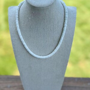 Op de afbeelding: Een witte kralenketting, gepresenteerd op een grijze mannequin. De ketting bestaat uit kleine, ronde kralen, die een delicaat en eenvoudig ontwerp vormen. De achtergrond is een wazig groen, wat duidt op een buitenomgeving.