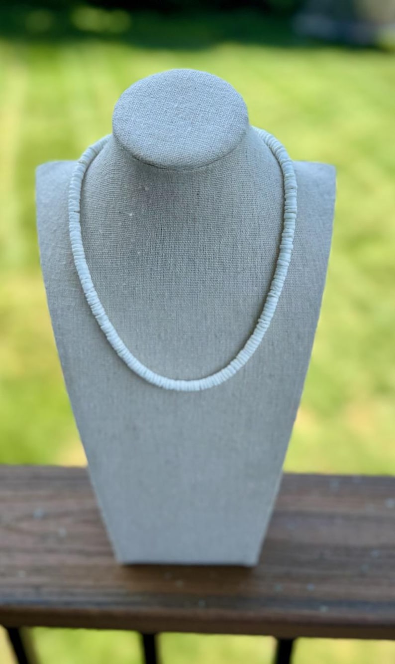 Op de afbeelding: Een witte kralenketting, gepresenteerd op een grijze mannequin. De ketting bestaat uit kleine, ronde kralen, die een delicaat en eenvoudig ontwerp vormen. De achtergrond is een wazig groen, wat duidt op een buitenomgeving.