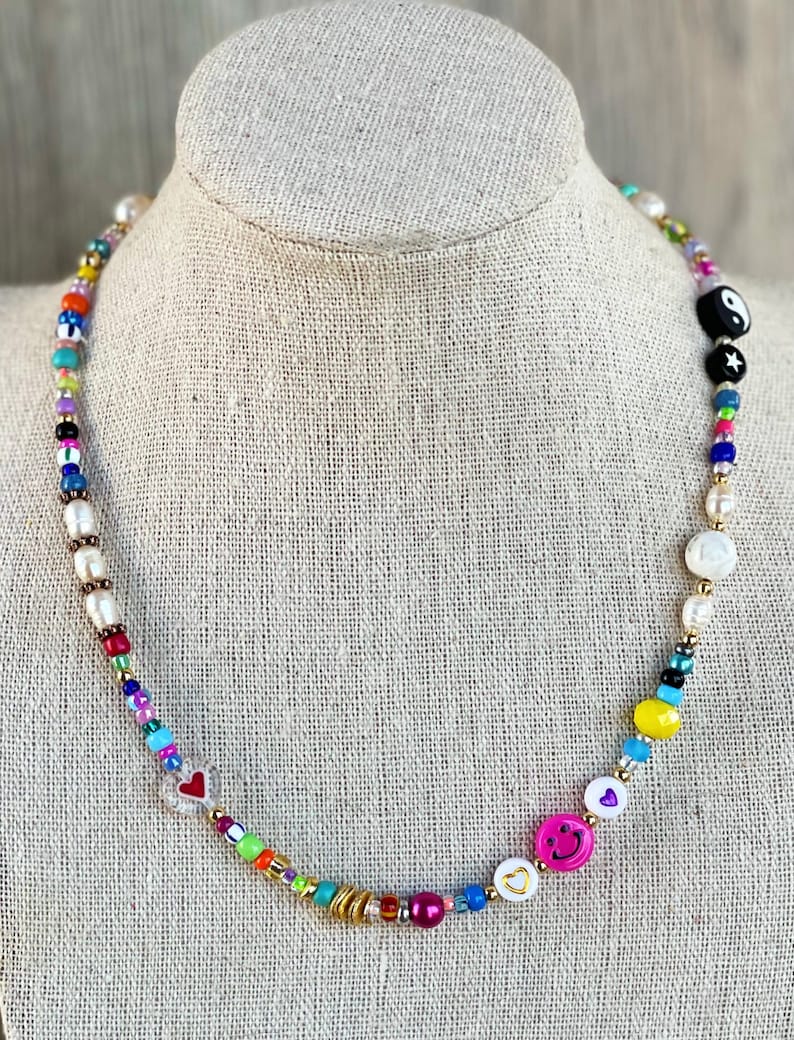 Op de afbeelding: Een kleurrijke kralenketting met verschillende bedels, waaronder harten, smiley-gezichten en een yin-yang symbool. De ketting is gemaakt met witte parels, gouden kralen en gekleurde glazen kralen.