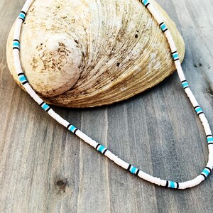 May include: White beaded necklace with turquoise and black accents. The necklace is displayed on a seashell against a wooden background. The necklace is a simple, yet stylish accessory.