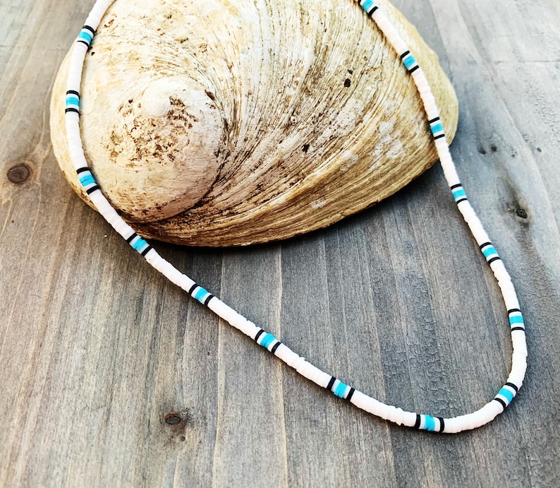 May include: White beaded necklace with turquoise and black accents. The necklace is displayed on a seashell against a wooden background. The necklace is a simple, yet stylish accessory.