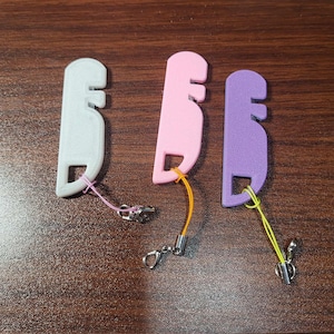May include: Three colorful, key-shaped accessories in white, pink, and purple. Each has a small loop with a lanyard and a metal clasp. The accessories are arranged on a dark brown wooden surface.