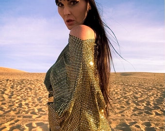 May include: A metallic gold, short-sleeved tunic featuring a dense, shimmering sequin or mesh texture. The garment has an off-the-shoulder neckline and a straight hemline. A matching gold fabric headband is worn across the forehead.