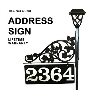 Puede incluir: Un letrero de direcci&oacute;n de metal negro con un dise&ntilde;o floral decorativo y una luz solar. El letrero muestra el n&uacute;mero 2364 en blanco sobre un fondo negro. El letrero est&aacute; montado en un poste de metal negro.