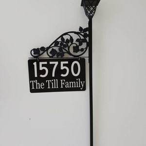 Puede incluir: Letrero de n&uacute;mero de casa de metal negro con un dise&ntilde;o floral decorativo y una luz solar. El letrero dice "15750 The Till Family".