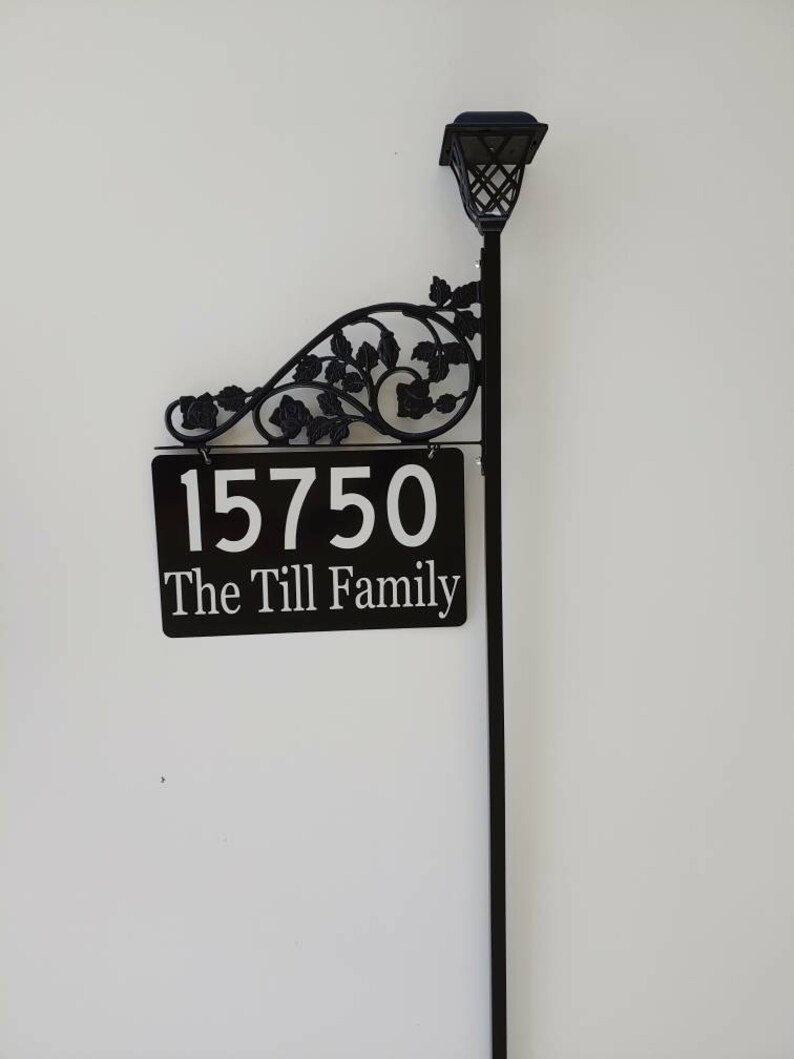 Puede incluir: Letrero de n&uacute;mero de casa de metal negro con un dise&ntilde;o floral decorativo y una luz solar. El letrero dice "15750 The Till Family".