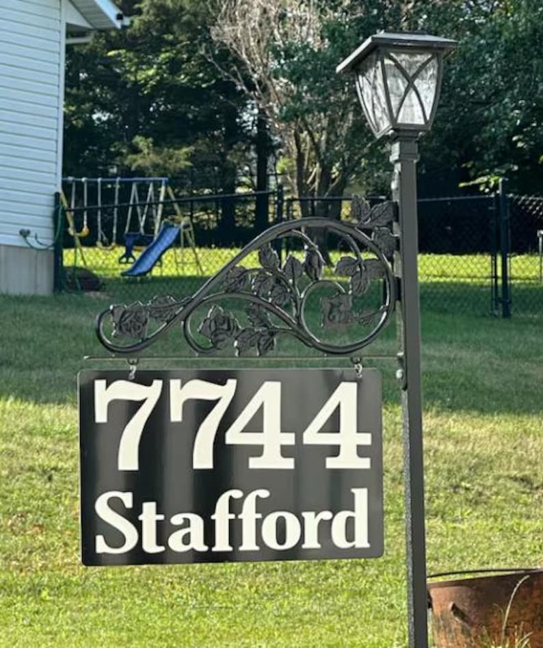 Puede incluir: Se&ntilde;al de n&uacute;mero de casa de metal negro con n&uacute;meros y texto blancos. La se&ntilde;al dice "7744 Stafford". La se&ntilde;al est&aacute; unida a un poste de metal negro con un dise&ntilde;o decorativo de rosas.
