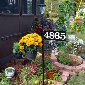 Puede incluir: Un letrero de n&uacute;mero de casa de metal negro con el n&uacute;mero 4865 sobre un fondo blanco. El letrero est&aacute; montado en un poste de metal negro con volutas decorativas. El letrero est&aacute; rodeado de una variedad de plantas y flores, incluyendo crisantemos amarillos, flores blancas y follaje verde.