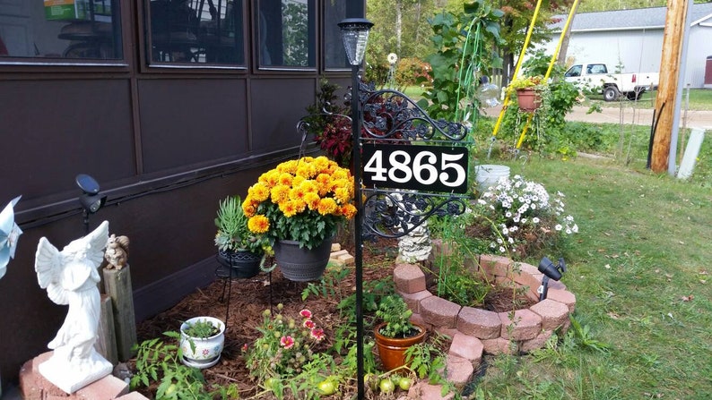 Puede incluir: Un letrero de n&uacute;mero de casa de metal negro con el n&uacute;mero 4865 sobre un fondo blanco. El letrero est&aacute; montado en un poste de metal negro con volutas decorativas. El letrero est&aacute; rodeado de una variedad de plantas y flores, incluyendo crisantemos amarillos, flores blancas y follaje verde.