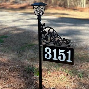 Puede incluir: Un letrero de n&uacute;mero de casa de metal negro con el n&uacute;mero 3151 sobre un fondo blanco. El letrero est&aacute; unido a un poste de metal negro con una luz solar en la parte superior.