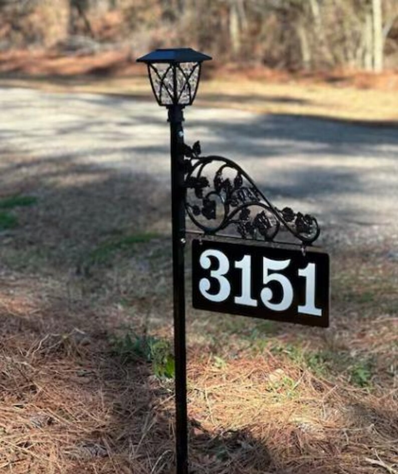 Puede incluir: Un letrero de n&uacute;mero de casa de metal negro con el n&uacute;mero 3151 sobre un fondo blanco. El letrero est&aacute; unido a un poste de metal negro con una luz solar en la parte superior.