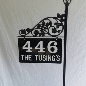 Puede incluir: Letrero de n&uacute;mero de casa de metal negro con un dise&ntilde;o floral decorativo y una luz solar. El letrero dice "446 The Tusing's".