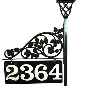 Puede incluir: Un letrero de n&uacute;mero de casa de metal negro con un dise&ntilde;o floral decorativo y una luz solar. El letrero muestra el n&uacute;mero 2364.