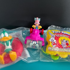 May include: Three Tiny Toon Adventures toys in plastic packaging. One features a green and yellow duck character on a red float. Another is a pink vehicle with a cartoon cat. The third is a yellow toy with the Tiny Toon Adventures logo.
