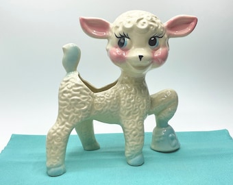 May include: A white ceramic lamb planter with blue and pink accents. The lamb has a curly, textured coat and large, blue eyes. The planter has a small opening in the back for planting.
