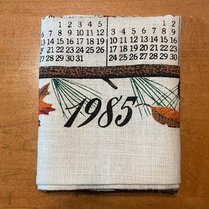 May include: Folded linen calendar towel featuring the year 1985. The towel has a calendar grid with dates and a brown border with leaves. The background is a light beige color.