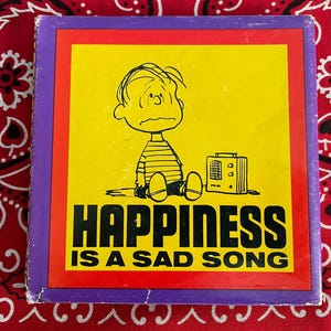May include: Square box with a yellow background and the text "HAPPINESS IS A SAD SONG". The image features a cartoon character sitting next to a radio. The box has a red and purple border.