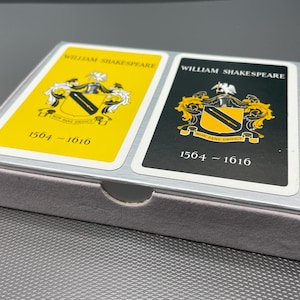 May include: A deck of playing cards in a silver box. The cards feature the name "WILLIAM SHAKESPEARE" and a crest design. One card has a yellow background, the other is black. The dates 1564-1616 and 1504-1610 are printed on the cards.