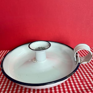 May include: White enamel candle holder with a handle. The candle holder has a circular base with a dark blue rim and a central cup for the candle. The handle is a curved loop. The background is red.