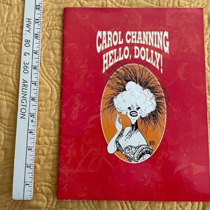 May include: A red program for the musical "Hello, Dolly!" starring Carol Channing. The program features a cartoon illustration of Carol Channing in a white wig and a red dress.