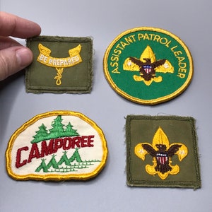 May include: Four vintage Boy Scout patches. The patches are green, yellow, and white with embroidered designs. One patch says "Assistant Patrol Leader" with a scout emblem. Another patch says "Be Prepared" with a scout emblem. A third patch says "Campore" with a design of trees and tents. The fourth patch has a scout emblem.