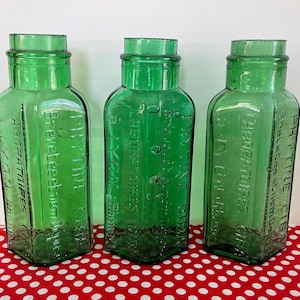 May include: Three vintage green glass bottles with embossed text. The bottles have a square base and a cylindrical neck. The bottles are displayed on a red and white polka dot surface.