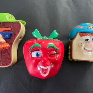 May include: Three vintage McDonald's Happy Meal toys. The toys are shaped like a steak wearing sunglasses, a red apple winking, and a sandwich with a blue hat. Each toy has a smiling face and is made of plastic.